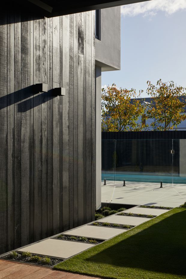 A contemporary residential exterior courtyard featuring dark vertical timber cladding, a projecting light-rendered upper volume, and a minimalist paved path of large rectangular stepping slabs through clipped lawn. A glass-fenced swimming pool and dark boundary fence sit beyond, with strong sunlight creating dramatic shadows across the facade and paving.