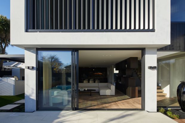 Contemporary two-storey residential facade with a broad covered opening leading into a living room, large sliding glass doors, white rendered walls, dark vertical upper screening, and warm timber flooring visible inside. A low landscaped garden and adjacent outdoor steps frame the modern home.