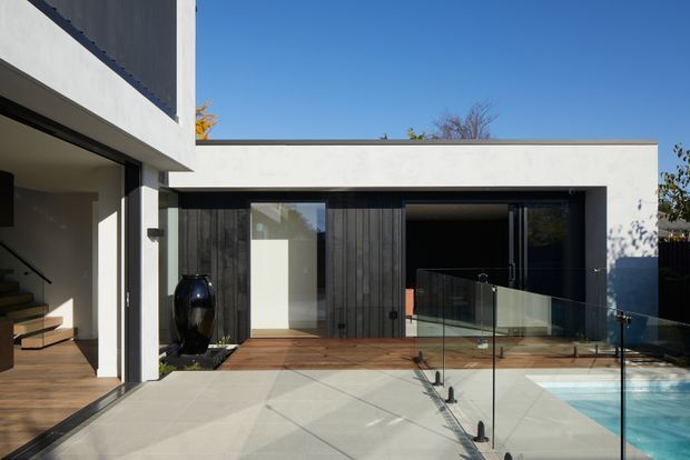 Exterior view of a contemporary flat-roof residence with white rendered walls, dark vertical timber cladding, expansive black-framed glazing, and a timber deck opening onto a swimming pool. A glazed balustrade separates the patio from the pool, while an entry stair and interior are visible at the left.
