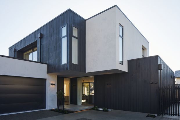 Contemporary multi-level residence viewed from the front, featuring a strong geometric composition of white rendered wall planes and dark vertical timber-clad volumes. The home includes a recessed entry, narrow vertical windows, an integrated double garage, a projecting upper storey, and a dark timber-clad side wing.