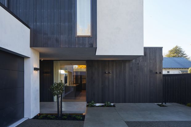 Contemporary residential facade viewed from the driveway, featuring dark vertical timber cladding, smooth white rendered walls, a projecting upper level, narrow vertical glazing, and a recessed entry walkway with simple landscaping. A dark metal roof and neighbouring residential context are visible in the background.
