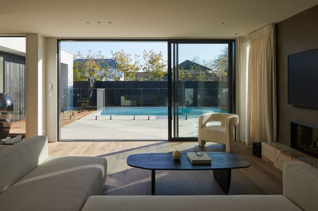 A refined contemporary living room with pale upholstered sofas, a dark oval coffee table, a wall-mounted television and a fireplace set into a dark feature wall. Full-height sliding glass doors open directly to a paved outdoor area with a swimming pool, while sheer curtains and warm timber flooring soften the neutral interior.
