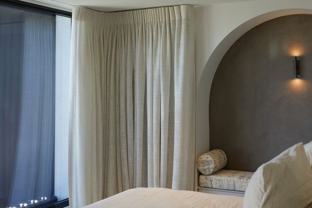 A softly lit bedroom corner with a large floor-to-ceiling window dressed in sheer and textured cream curtains. The bed is partially visible in the foreground, while a built-in arched alcove with a textured taupe plaster finish contains a cushioned bench and a cylindrical woven cushion. A slim wall sconce provides warm accent lighting.