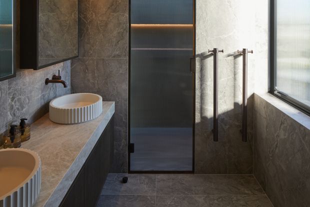 A compact contemporary bathroom featuring a double vanity with distinctive fluted round vessel basins, a stone-look benchtop and wall finish, dark cabinetry, black-framed ribbed-glass shower enclosure, and tall brushed metal tapware. Natural light enters through a large window, creating a refined, warm-toned interior.