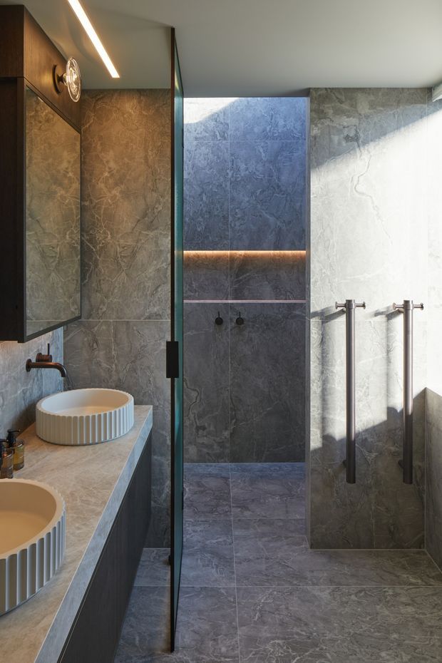 A refined contemporary bathroom featuring a walk-in shower enclosed by a frameless glass screen, dark grey veined stone-look wall and floor tiles, a pale stone vanity with two round fluted vessel basins, dark timber cabinetry, brushed metal tapware, and integrated warm linear lighting. Strong natural light creates a high-contrast, luxurious atmosphere.