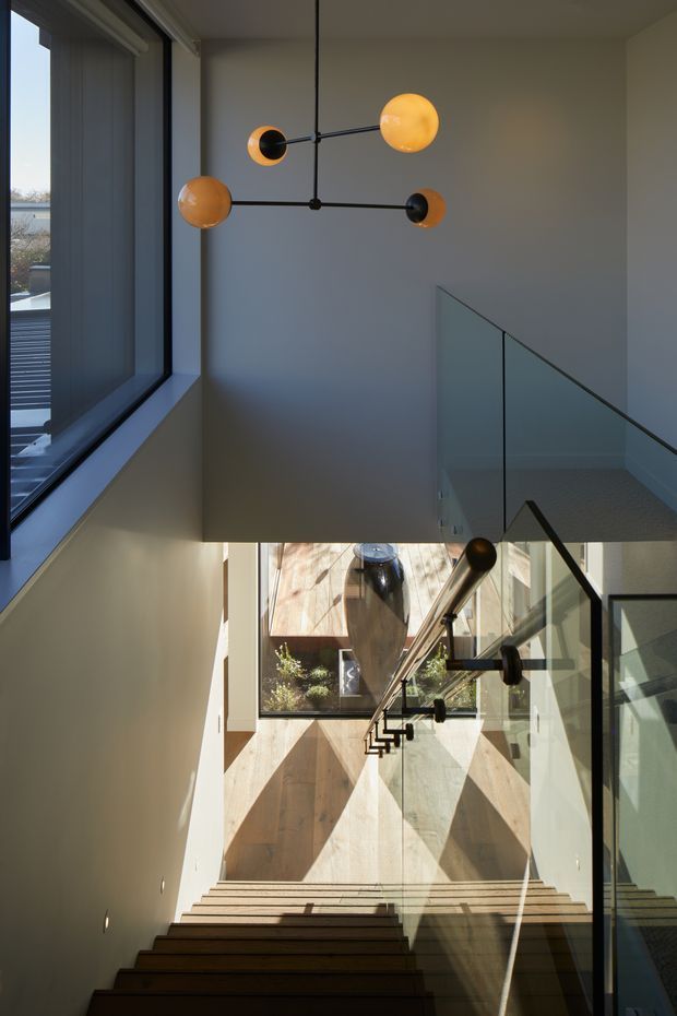 A contemporary stairwell viewed from below and above, featuring a timber staircase with black metal handrails, clear glass balustrades, pale plaster walls, and large windows bringing in strong natural light. A sculptural black-and-amber globe pendant hangs in the upper stair void, while a large dark ceramic vessel and timber-clad wall create a warm focal point at the landing.