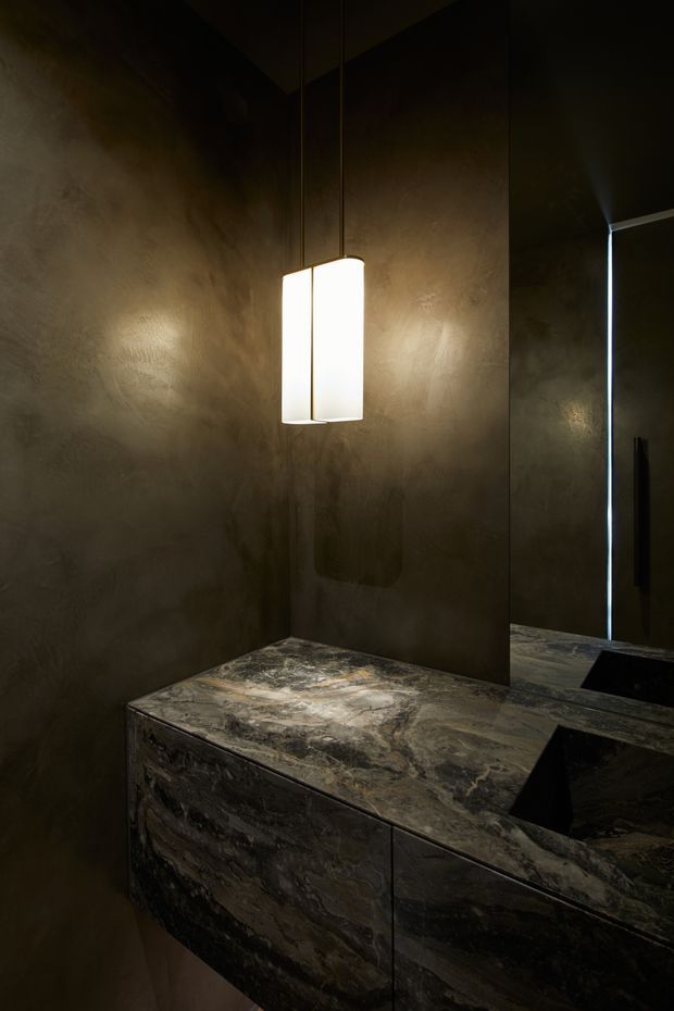 Moody, luxurious bathroom vanity with dark textured plaster walls, a dramatic veined natural-stone benchtop and integrated dark cabinetry. A suspended rectangular pendant light illuminates the compact vanity, with a large mirror and two dark angular basins visible.