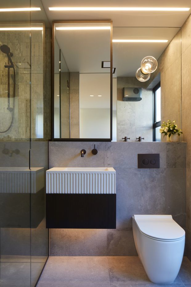 A contemporary bathroom with a walk-in glass shower, wall-mounted ribbed vanity, large black-framed mirror, wall-hung toilet, concrete-look tiled walls and floor, and integrated linear ceiling lighting. Black fixtures and a sculptural wall light provide contrast against the warm grey finishes.