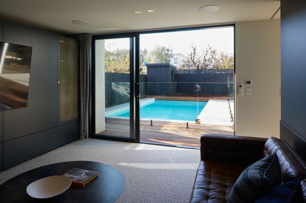 A contemporary living room with dark built-in cabinetry and a wall-mounted television, plush light carpet, a round black coffee table, and a dark leather sofa. Large glazed sliding doors open directly to a timber deck and bright backyard swimming pool, creating a strong indoor–outdoor connection.
