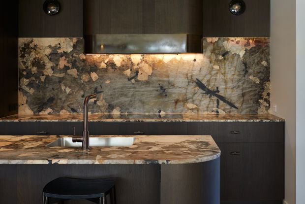 A dark, sophisticated contemporary kitchen featuring extensive dramatic natural stone on the island benchtop and splashback, dark wood cabinetry, an undermount sink with tall curved tap, integrated cooktop and hood, and subtle warm under-cabinet lighting. A black bar stool sits at the island.