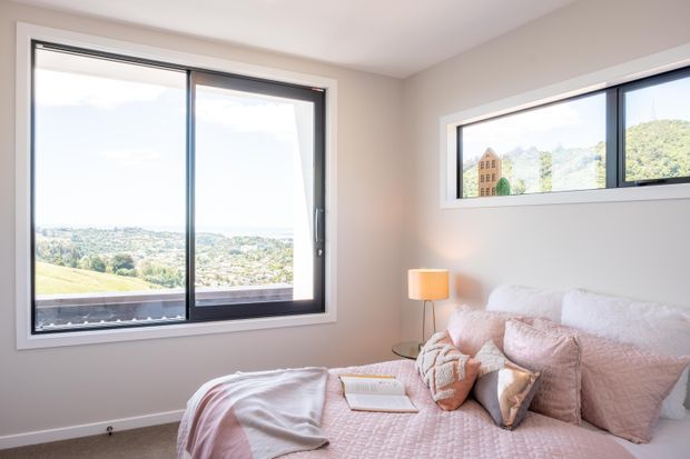 Bright, compact bedroom with a large black-framed sliding window and a narrow horizontal window, offering elevated views over a leafy residential landscape. The room has pale warm-grey walls, carpeted flooring, and a blush pink quilted bed with coordinated cushions and a small bedside lamp.