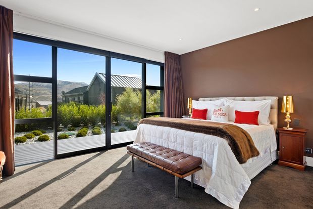 A refined bedroom with a large upholstered bed, white quilted bedding, red accent cushions, and a brown throw. A deep brown feature wall is paired with full-height black-framed glazing and burgundy curtains, opening to a landscaped garden and mountain views. A tufted leather bench sits at the foot of the bed, with warm bedside lamps and dark timber bedside tables.