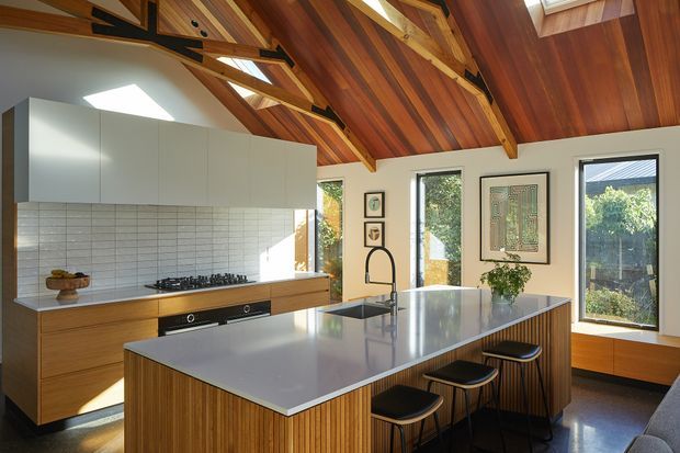 A bright contemporary kitchen with a large central island, integrated cooktop and oven, pale cabinetry, white tiled splashback, and warm timber island and lower cabinetry. The room is defined by a dramatic pitched timber-lined ceiling with exposed structural beams, roof glazing, and clerestory-like windows overlooking greenery. Bar seating, framed artwork, and indoor planting add a refined residential character.