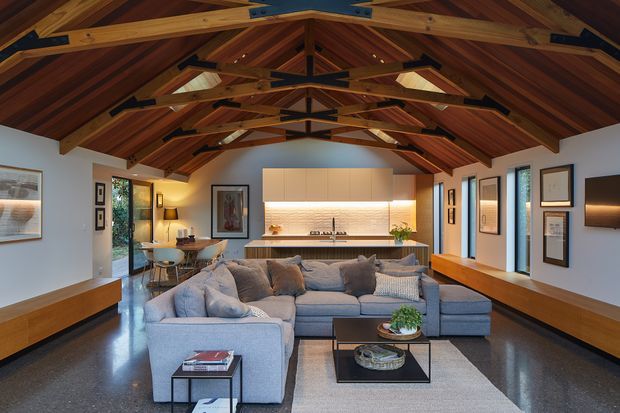 A spacious open-plan living room with a large sectional sofa, polished dark flooring, and extensive custom built-in cabinetry. The room features a dramatic pitched timber ceiling with exposed structural beams and rafters, skylights, and warm integrated lighting. A contemporary white kitchen and dining area sit at the far end, with large windows and glazed doors bringing in natural light. Framed artwork, timber joinery, and layered neutral furnishings create a refined, warm interior.