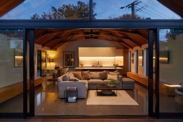 View through fully open dark-framed glazed doors into a warm, open-plan living room with a prominent exposed timber vaulted roof structure. The room contains a large sectional sofa, rug, coffee table, dining area, and a linear kitchen with pale cabinetry and illuminated textured backsplash. Polished concrete or terrazzo-like flooring, timber wall and ceiling elements, and extensive glazing create a refined indoor-outdoor connection. Exterior trees and utility lines are visible through the glazing.