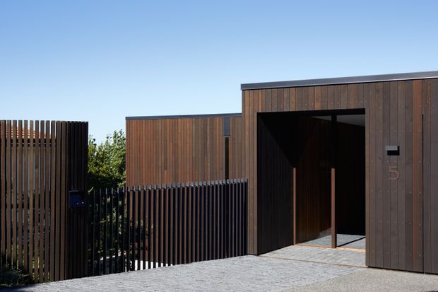 Contemporary dark timber-clad residential entrance viewed from the driveway, featuring a flat-roofed stepped facade, tall vertical slatted fencing, a recessed entry portal, and grey paved surfaces under clear daylight.