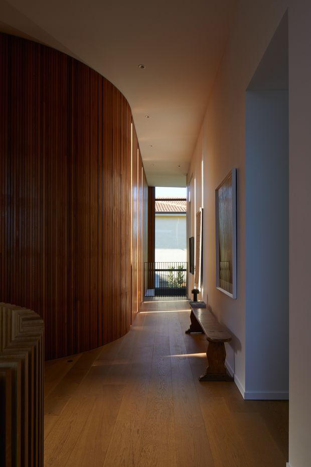 A long, narrow residential hallway with warm timber flooring and a dramatic curved wall clad in vertical dark wood slats. The corridor has smooth off-white walls and ceiling, recessed downlights, framed artwork, and a simple timber console bench. Natural light enters from an opening at the far end, where a small balcony railing and terracotta-tiled roof are visible, creating a warm, refined architectural interior.
