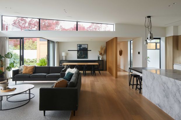 A bright, open-plan contemporary home interior combining a living room, dining area and kitchen. The room features expansive glazing, warm timber flooring, a black-framed window and door system, a vertical timber slat partition, a dark dining table, charcoal sofas, and a large white-grey veined stone kitchen island. A sculptural pendant hangs above the island, while indoor plants and neutral furnishings add warmth.