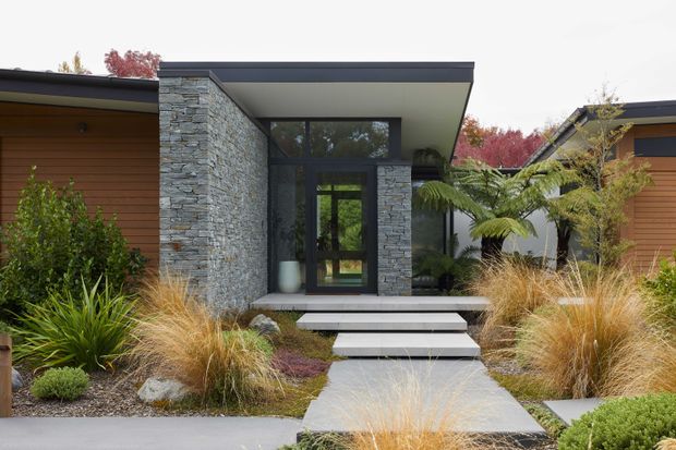 Contemporary residential entry facade with a strong central portal framed in stacked natural stone, dark metal-framed glazing, a broad flat roof canopy, and warm horizontal timber cladding to the wings. Wide concrete steps and a path lead through a layered native-style garden with ornamental grasses, ferns, shrubs, and textured gravel.