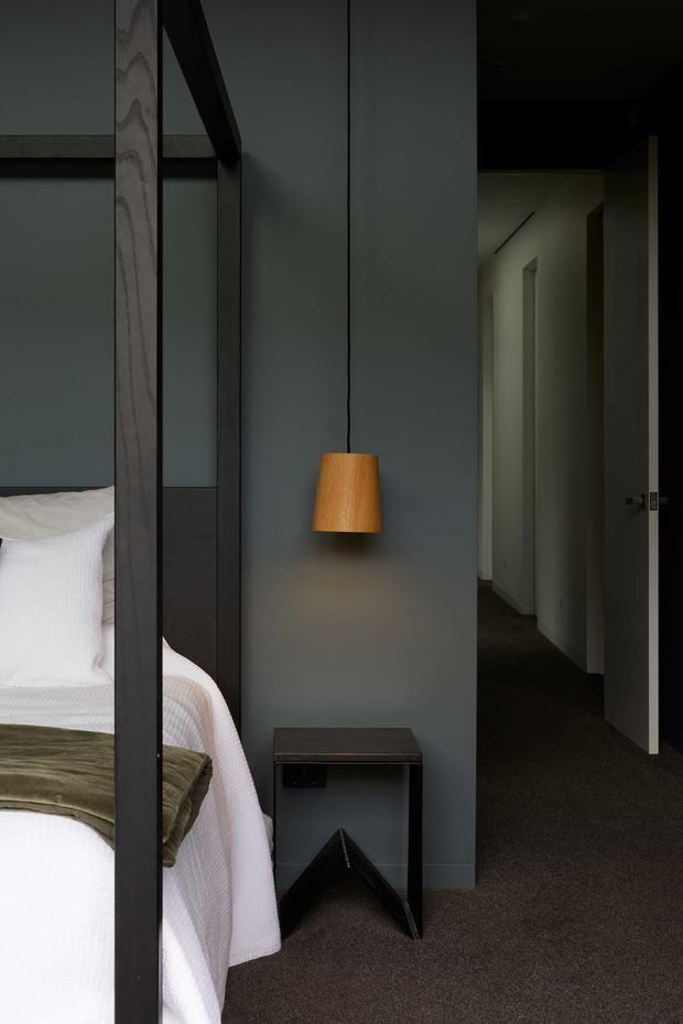 A moody, minimalist bedroom viewed beside a dark-framed four-poster bed. The room has charcoal-grey walls, white textured bedding with an olive-green throw, dark carpet, a small black bedside table, and a suspended warm wood pendant light. A hallway with open doors is visible beyond the bedroom.