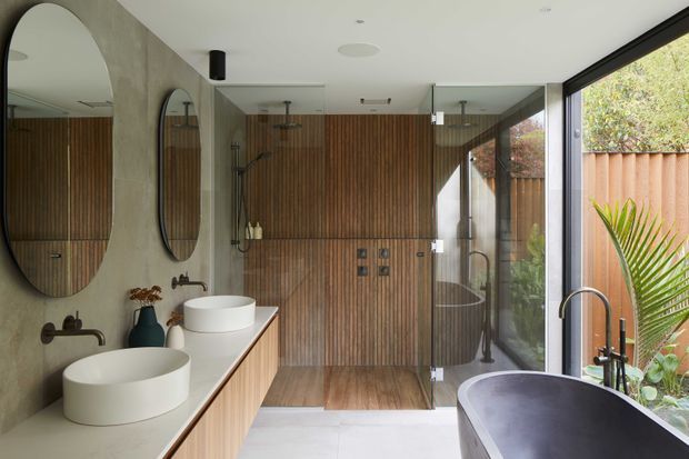 Contemporary bathroom with double vessel basins on a long timber vanity, brushed dark-metal wall-mounted taps, a freestanding dark bathtub, and a walk-in glass shower. Vertical timber wall panelling contrasts with a pale stone-look vanity and light floor tiles. Full-height glazing opens to a timber-fenced garden, creating a bright, refined space.