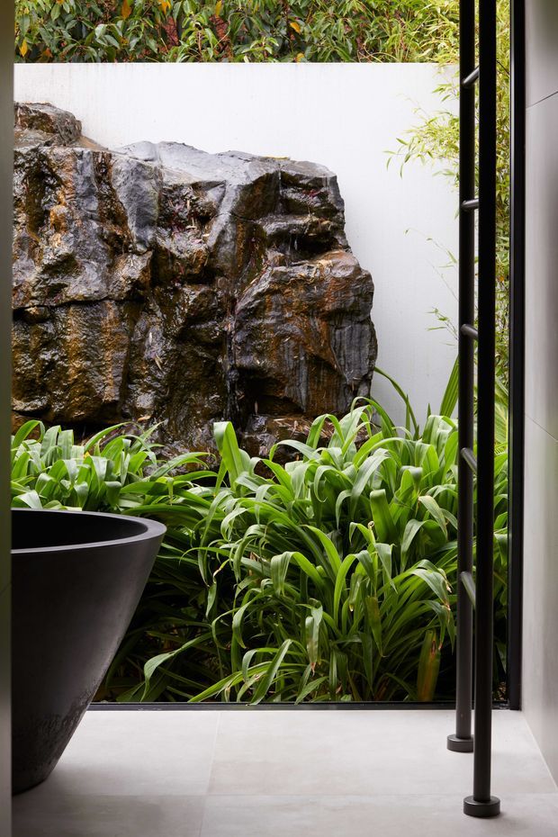 A refined indoor-outdoor view from a minimalist interior toward a lush planted courtyard. A large rugged natural-rock water feature forms the backdrop, surrounded by dense green strap-leaf planting. The space has pale smooth tiled flooring, a white rendered wall, and slim black-framed floor-to-ceiling glazing or an open framed threshold. A black freestanding planter is visible in the foreground, with dark vertical metal supports adding a contemporary architectural detail.