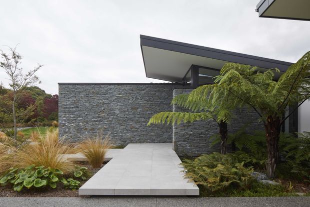 Exterior view of a contemporary low-profile building approached by a broad pale paved walkway. The facade features extensive horizontally layered grey stone cladding, dark fascia and roof edges, and large projecting flat roof planes. Lush landscaped planting, including tree ferns, grasses, and hostas, frames the entrance and complements the restrained architectural palette.