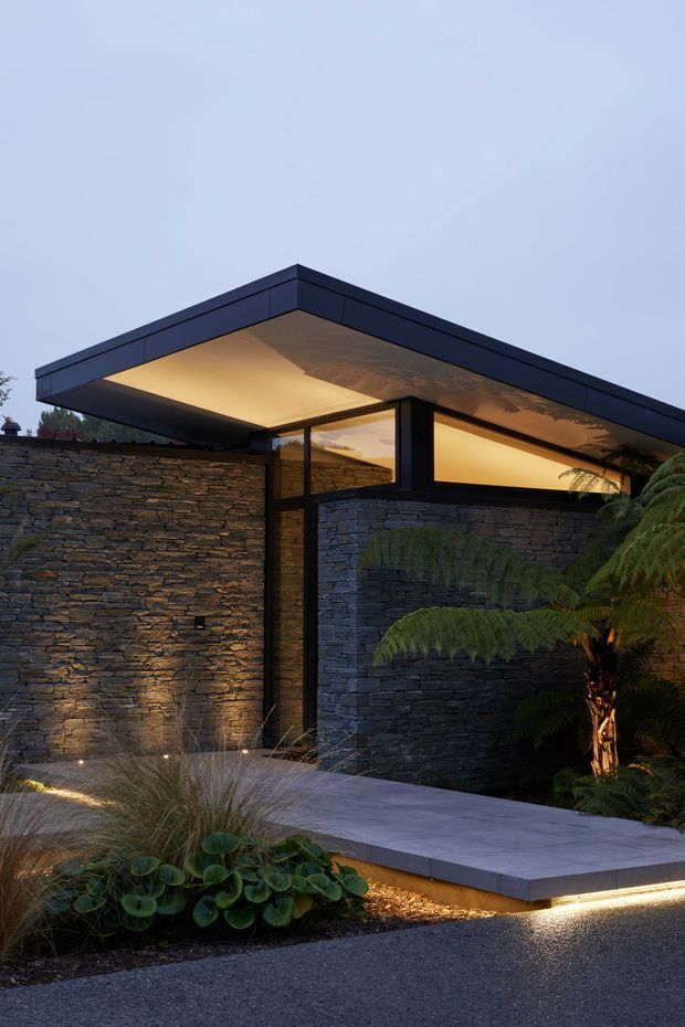 A dusk exterior view of a contemporary low-profile entrance or courtyard. Dark stacked-stone walls form a sheltered passage beneath a broad flat roof with deep cantilevered eaves and warm recessed lighting. Black-framed clerestory glazing sits beneath the roofline, while a pale paved path and layered native-style planting, including tree ferns and grasses, frame the approach.
