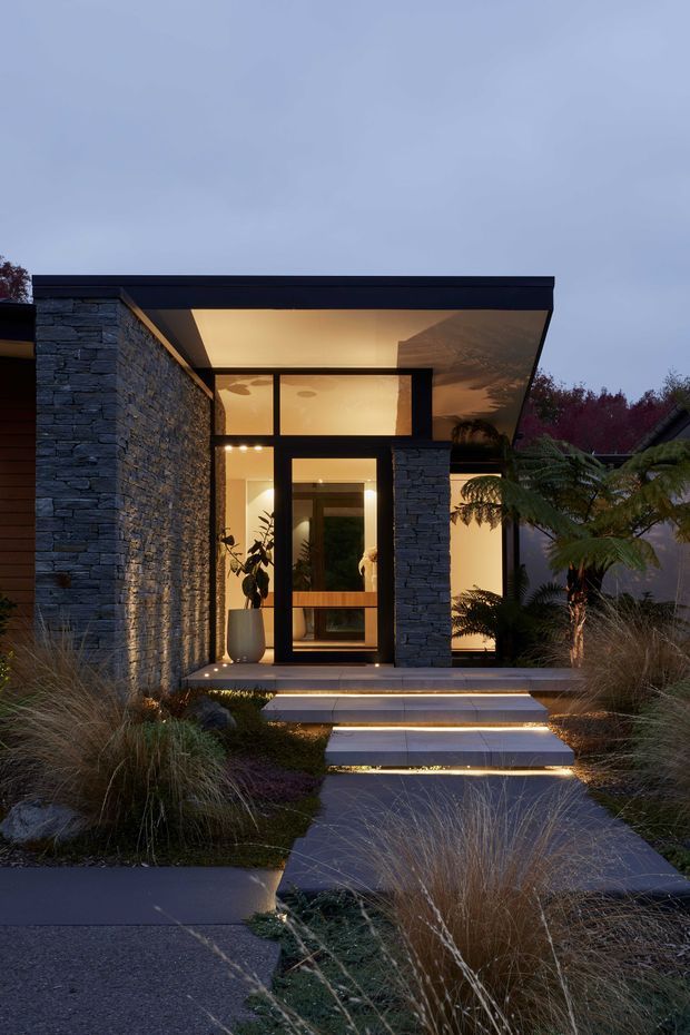Dusk exterior view of a contemporary single-storey home entrance. A flat-roofed porch projects over a glazed entry, framed by dark metal and rough stacked-stone walls. Broad illuminated steps lead through a landscaped front garden with ornamental grasses and sculptural planting. Warm interior lighting contrasts with the cool blue evening light.