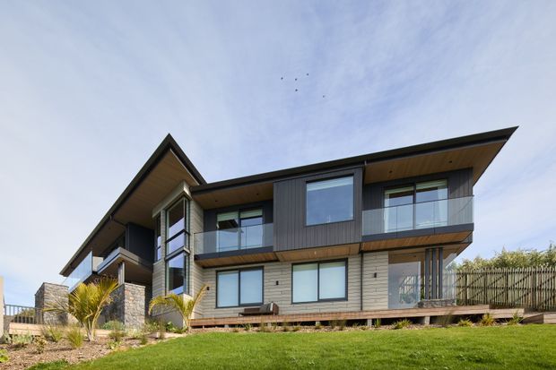 Exterior view of a contemporary multi-level residence with strong horizontal roof planes, projecting eaves, extensive glazing, balconies, vertical dark cladding, pale masonry walls, and stone-clad elements. The home sits within a landscaped suburban site with a lawn and timber deck.