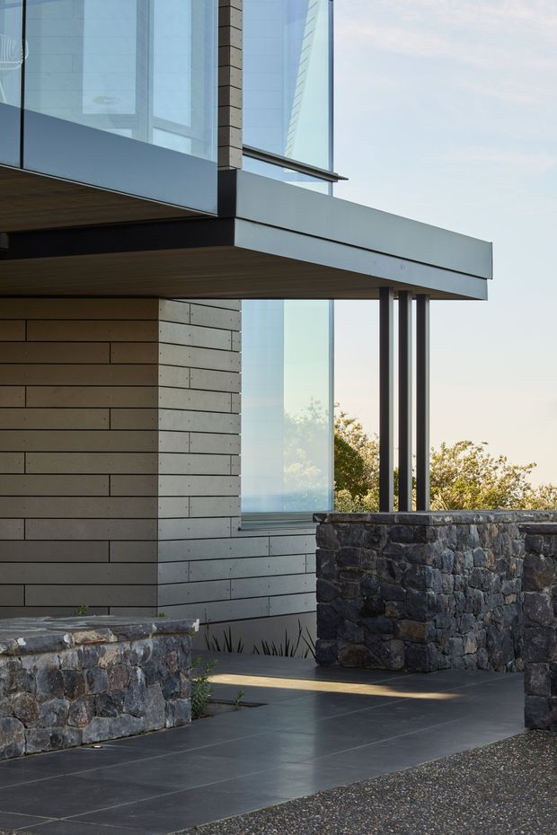 Close-up exterior view of a contemporary multi-level building with extensive floor-to-ceiling glazing, projecting flat roof and balcony forms, dark metal framing, horizontal light-toned cladding, and substantial dark natural-stone boundary walls. A paved entry path and terrace occupy the foreground, with landscaping visible beyond the glazing.