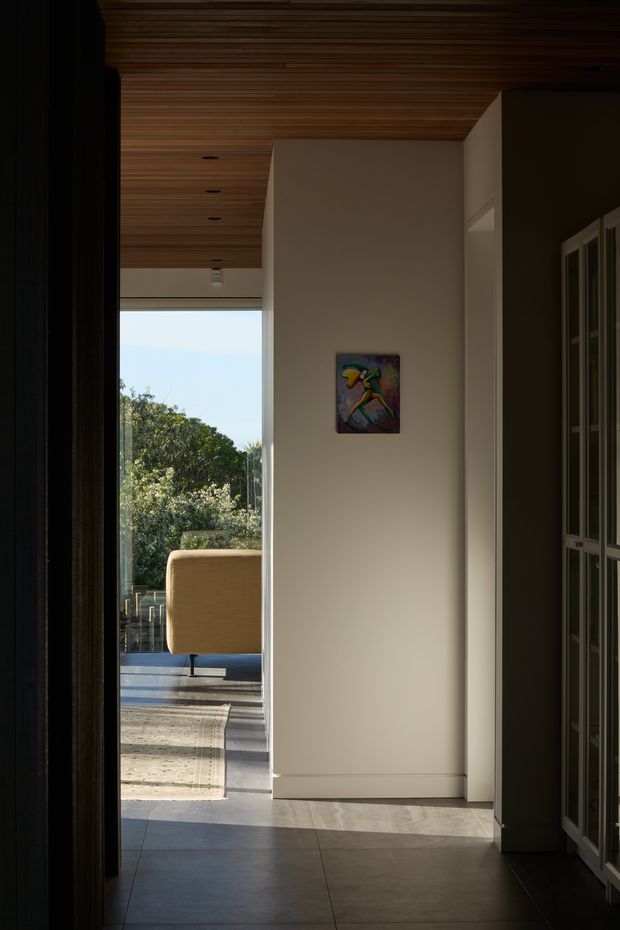 A dimly lit interior passage or entry hall leading toward a bright outdoor terrace. The space features a warm horizontal timber-lined ceiling, smooth off-white walls, large grey floor tiles, and tall dark built-in cabinetry along the right side. A small colourful artwork hangs on the central wall, while an upholstered outdoor chair and greenery are visible beyond the opening. The image has a refined, modern architectural character with strong contrast between the shadowed hall and sunlit exterior.