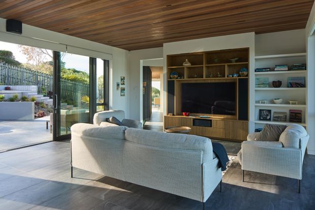 Contemporary living room with a large light-coloured sofa and armchair arranged around a built-in timber media wall. The room has extensive glazing opening to an outdoor terrace, white walls, dark tiled flooring, open display shelving, and a warm timber-lined ceiling.