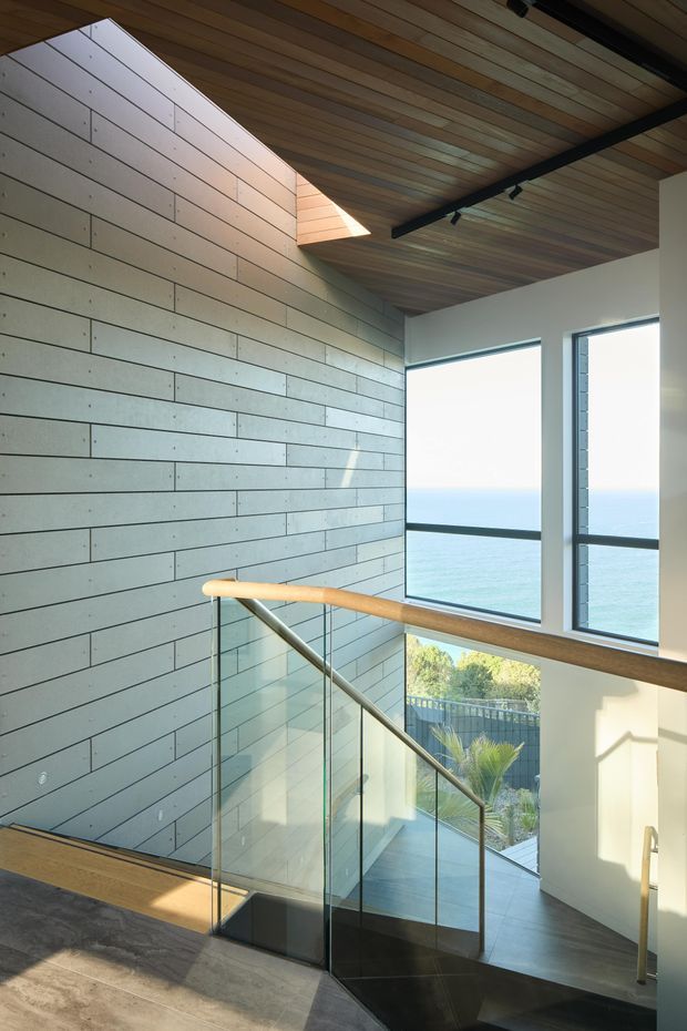 A contemporary stairway and landing with a sculptural timber-lined ceiling, a narrow skylight, and a long horizontal wall clad in pale rectangular panels. Glass balustrades with timber handrails border dark stairs, while large windows provide expansive ocean views and abundant natural light. Black track lighting contrasts with the warm timber ceiling.
