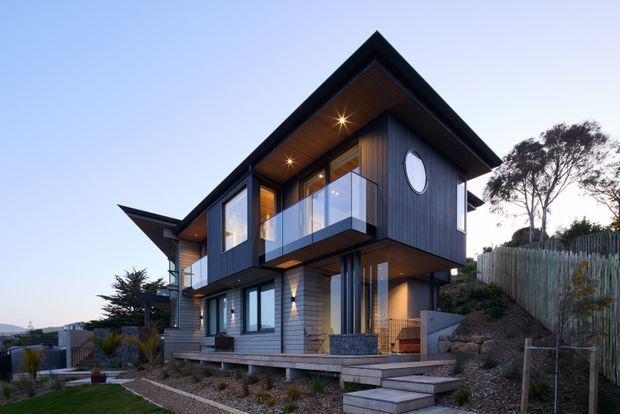 Exterior architectural photograph of a contemporary multi-storey house on a sloped suburban site. The home features dark vertical cladding, extensive glazing, projecting balconies with clear glass balustrades, a steep gabled roof with timber-lined soffits, and an elevated entry approached by stepped timber decking. Landscaping, retaining elements, and neighboring homes are visible around the building.