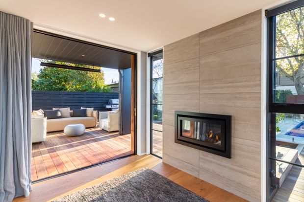 Bright contemporary living room with a built-in linear fireplace set into a full-height light timber-look feature wall. Warm timber flooring continues through large glazed openings to a covered outdoor deck and lounge area, with additional views toward a swimming pool. Grey curtains and a textured rug add softness to the refined, indoor-outdoor space.