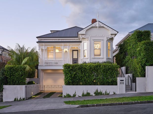 Street-facing exterior view of a renovated single-storey heritage-style residence with ornate white timber detailing, a prominent gabled front bay, decorative verandah, corrugated metal roofing, enclosed garage beneath the raised house, manicured hedges, front fencing and driveway. The dwelling sits in a dense suburban streetscape with neighbouring homes visible on either side.