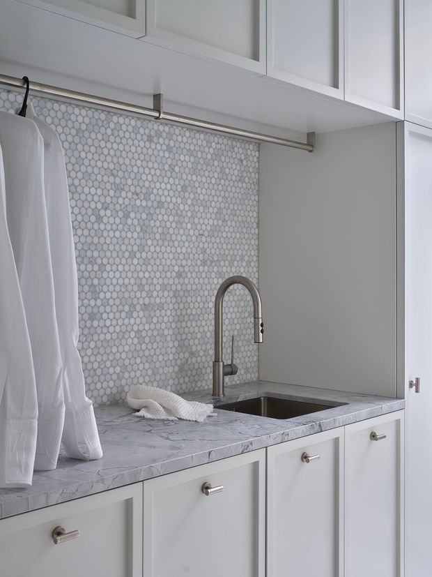 Close-up of a bright, compact laundry room with white built-in cabinetry, a stainless steel hanging rail, marble-look benchtop, small undermount sink, brushed metal gooseneck tap, and a white and grey penny-round mosaic tile splashback. White garments hang from the rail.