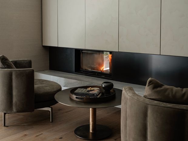 A refined contemporary living room centred on a built-in linear fireplace set into a dark feature wall beneath pale integrated cabinetry. Two rounded upholstered lounge chairs and a circular coffee table sit over warm timber flooring, creating an intimate, sophisticated seating area.