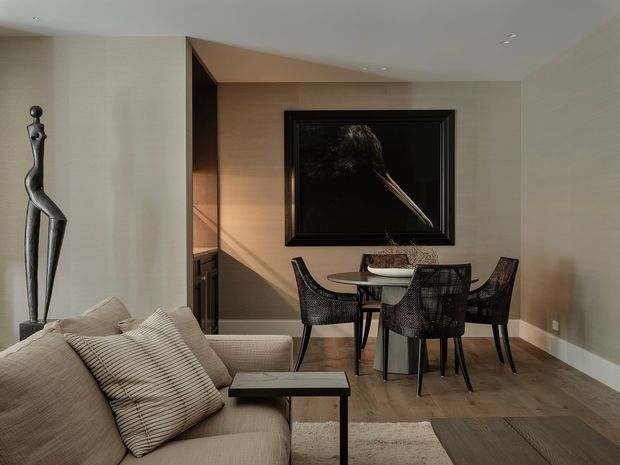 A refined contemporary dining and living area with a neutral palette. The room features a beige upholstered sofa with striped cushions, a round dining table with dark woven dining chairs, a slim black side table, light timber flooring, recessed ceiling lighting, and a large dark framed artwork. A sculptural figurine stands beside the sofa, while built-in dark cabinetry is visible in a recessed alcove.