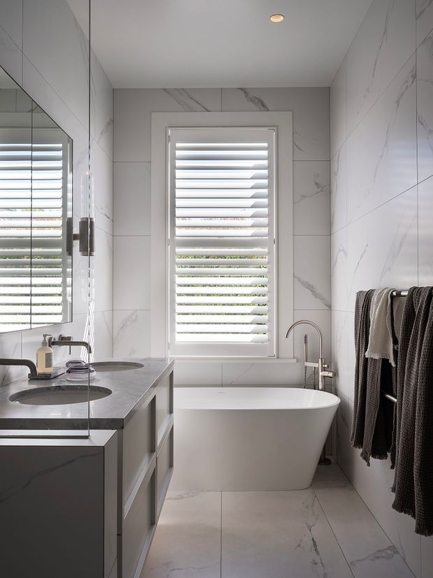 Bright, compact contemporary bathroom with large white marble-look porcelain tiles, a freestanding white bathtub beneath a shuttered window, a wall-mounted or compact double vanity with dark stone benchtop, mirrored cabinetry, brushed metal tapware, and a towel rail with dark textured towels. The space is clean and refined with strong natural light and a neutral palette.