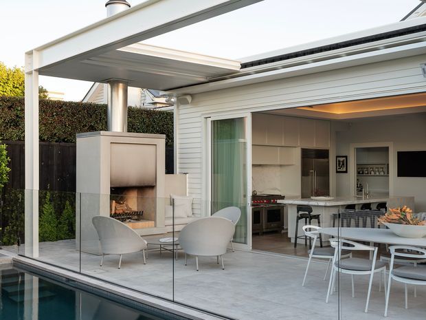 A bright contemporary outdoor entertaining area beside a swimming pool, with a glass balustrade, pale tiled terrace, built-in outdoor fireplace, sculptural lounge chairs, and a covered open-plan kitchen and dining area beyond. The architecture uses white horizontal cladding, broad roof overhangs, and large sliding openings to connect the interior and exterior.