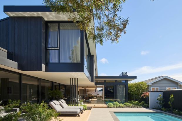 Contemporary multi-level residential exterior with dark vertical cladding, extensive glazing, projecting upper-floor volumes, and a flat roofline. The view includes a landscaped outdoor living area with loungers and a rectangular swimming pool, plus a partially visible open-plan interior beneath the cantilevered upper level. A neighbouring gabled house is visible beyond the property.