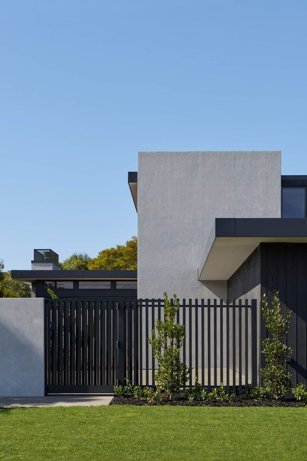 Street-facing view of a contemporary multi-level residence with a crisp rendered concrete-grey facade, dark vertical timber or metal cladding, black-framed glazing, and a projecting flat-roofed upper volume. A tall black slatted privacy gate and fence define the front boundary, with simple landscaped planting and a manicured lawn in the foreground under a clear blue sky.