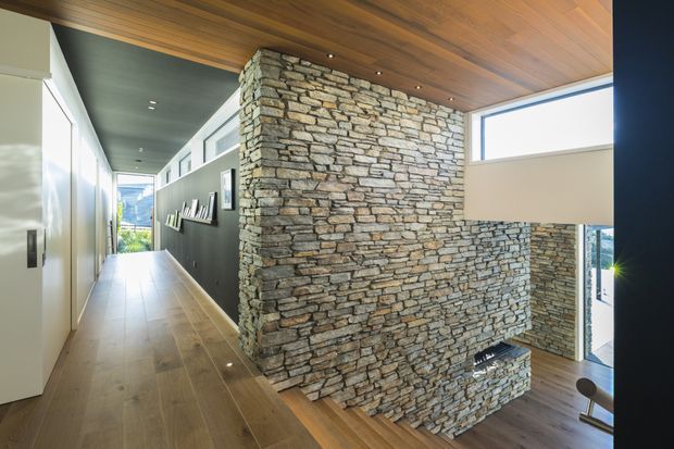 A contemporary interior hallway with a long linear circulation space, pale timber flooring, and a dramatic exposed stacked-stone feature wall forming a central vertical element. The ceiling is clad in warm timber boards with recessed downlights, while dark charcoal walls, crisp white built-in cabinetry, large windows, and a high horizontal clerestory bring contrast and natural light. A glimpse of an adjacent lower level is visible beyond the stone wall.