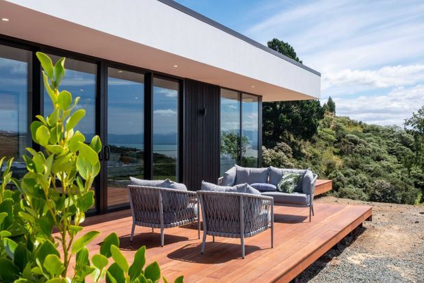 Contemporary flat-roofed home viewed from an elevated timber deck, with extensive black-framed glazing, dark vertical cladding, and a covered outdoor seating area overlooking a lush coastal landscape.