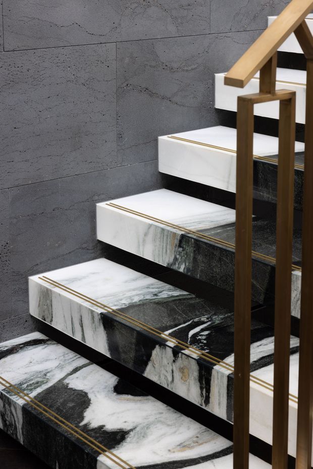 Close-up interior view of a sculptural staircase with floating white and dark veined natural-stone treads, set against a dark grey stone-tiled wall. Thin brass or gold inlays accent the tread edges, while a warm timber handrail and dark bronze-toned balustrade frame the stair.
