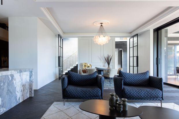 A refined contemporary living room with two dark upholstered armchairs, a sculptural round coffee table, a marble-clad fireplace, and a dining area beyond. Black-framed glazed doors, white panelled walls, ornate ceiling mouldings, and a statement clustered pendant create a polished, high-contrast interior.