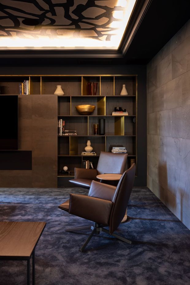 A moody, high-end media or living room with dark built-in shelving, bronze-toned detailing, a wall-mounted television, two leather lounge chairs, and a low timber coffee table. The room features a dramatic patterned illuminated ceiling, dark textured wall finishes, and a blue-grey patterned carpet, creating a luxurious contemporary atmosphere.