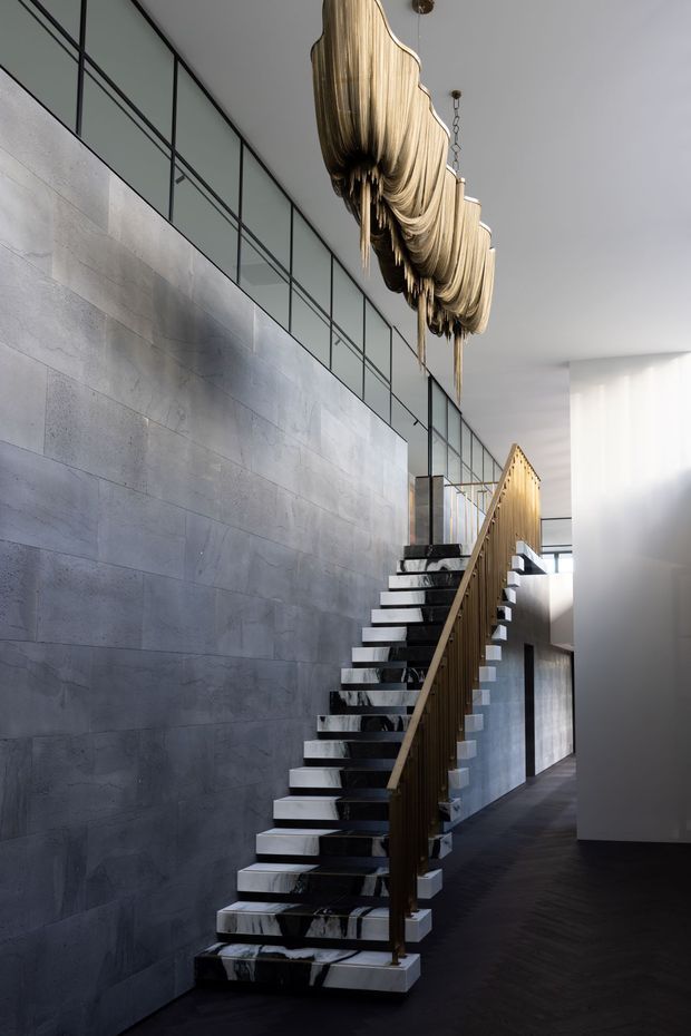 A dramatic contemporary interior stairway with floating marble treads, a slender brass-toned balustrade, dark flooring, and a large sculptural draped pendant light. The staircase is set against a pale grey stone-clad wall with a glazed upper-level balustrade, creating a refined, gallery-like atmosphere.